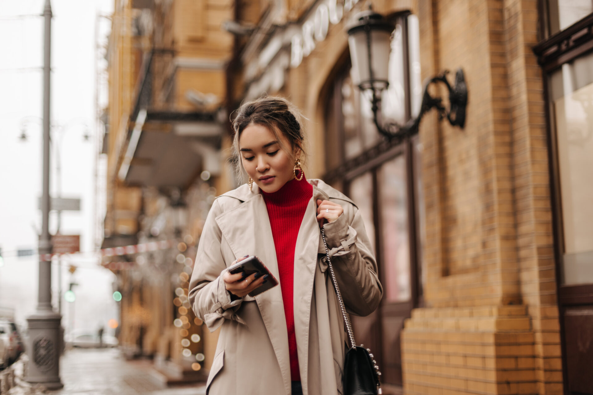 Stylish Asian lady in beige coat, red top and cross-body bag walks around city, holding smartphone